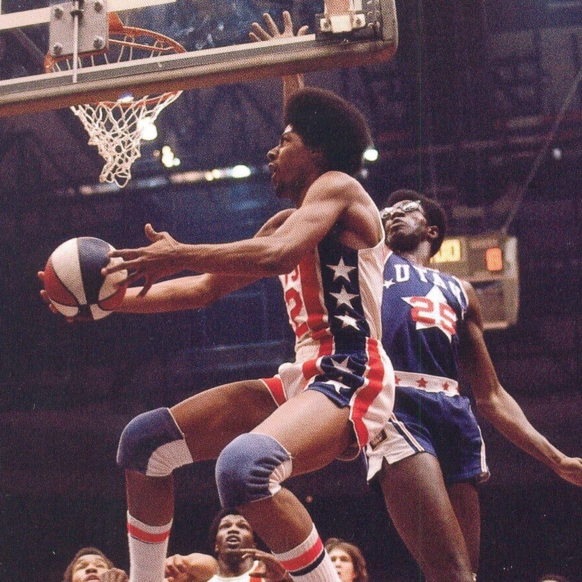 Julius Erving of the New York Nets maneuvers under the basket for a reverse layup during an ABA game, with a defender from the Utah Stars closely contesting. The scene highlights Erving's athleticism and creativity on the court.